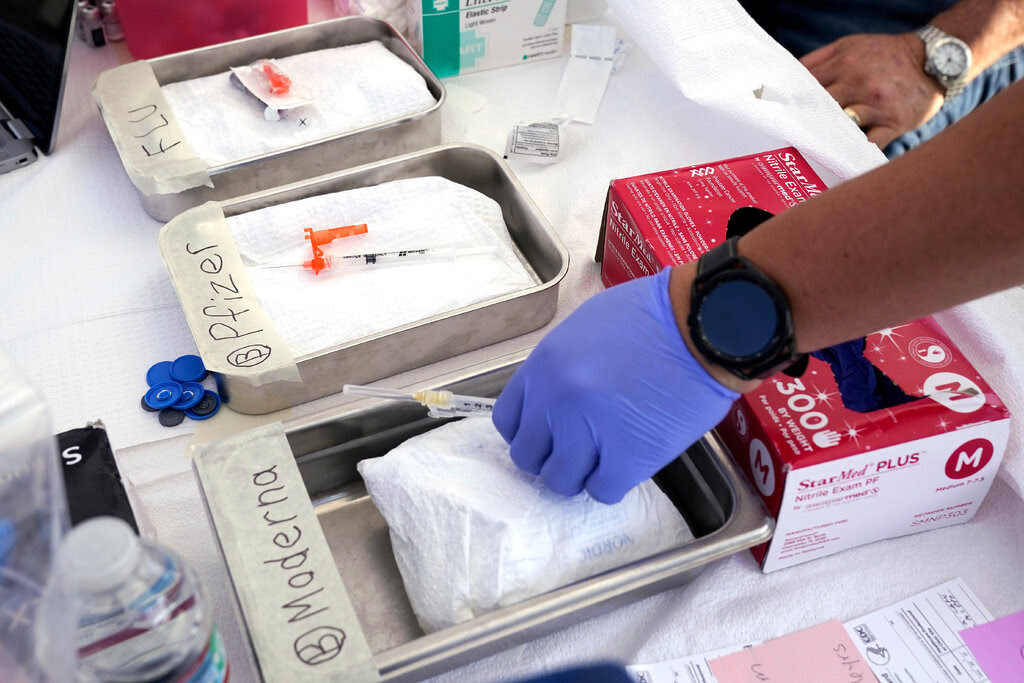 A purple-gloved hand holds a syringe in a silver bin labeled “Moderna.” Two other bins with syringes lie next to it, one reading “Pfizer” and the other reading “Flu.”