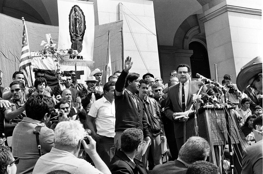 A man stand with his hand raised surrounded by a group in front of microphones.