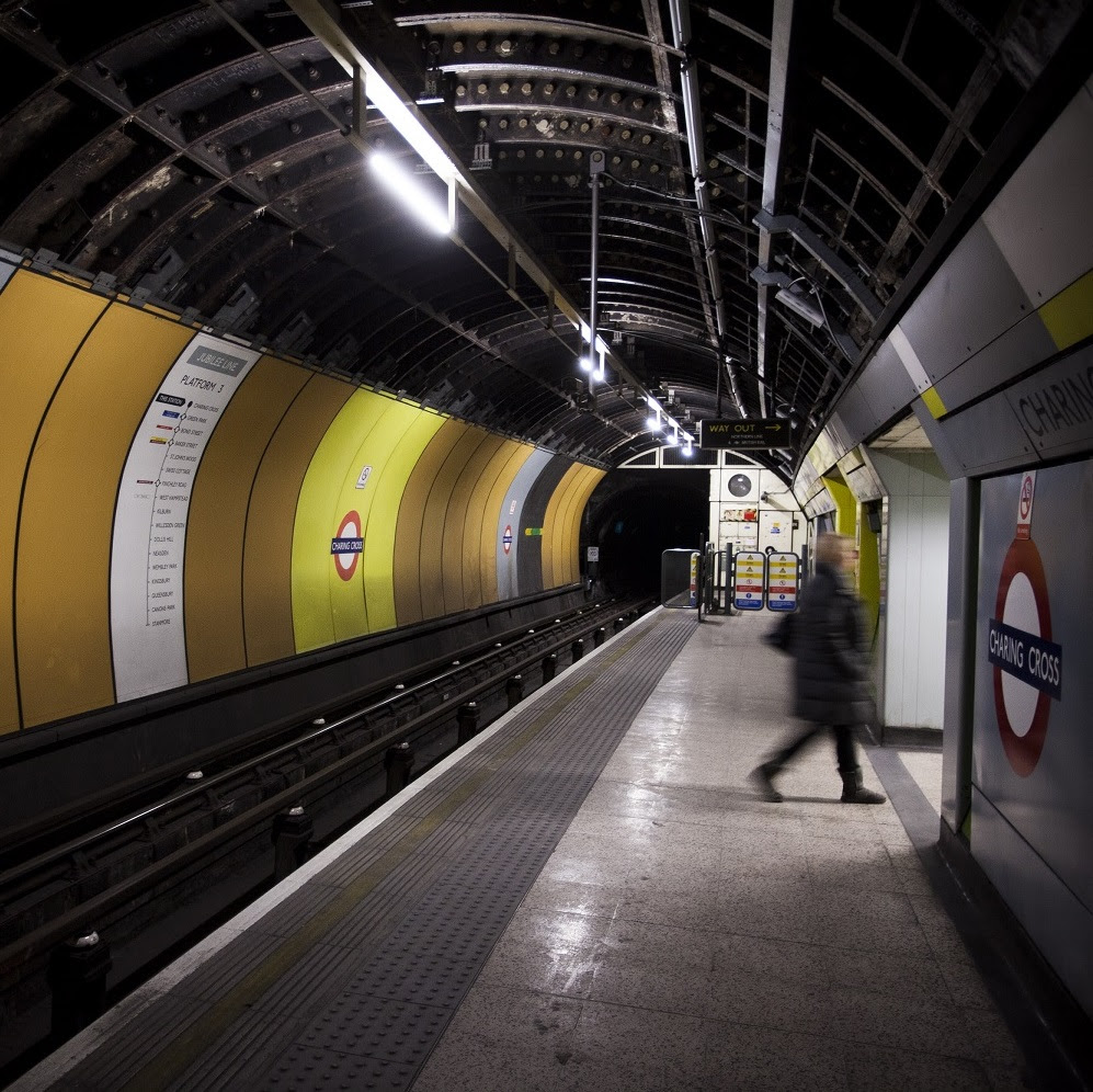 A blurred human figure walks off an empty Underground station platform with yellow and orange walls and Charing Cross roundels