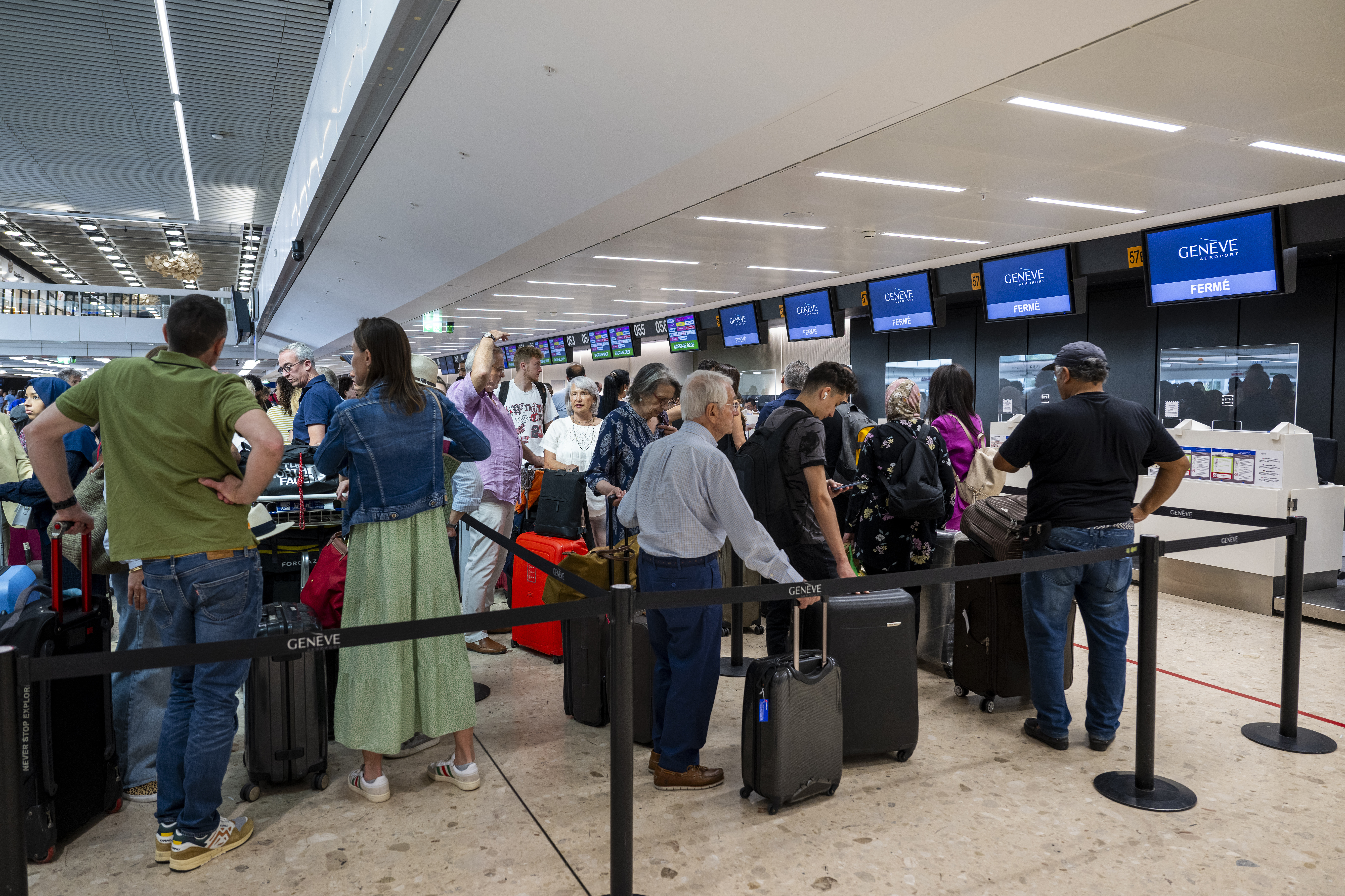 Des passagers attendent avec leurs bagages devant les guichets d'embarquement du terminal principal de l'aeroport international de Geneve, tandis que le syndicat et des employes de l'aeroport de Geneve (AIG) qui manifestent a l'exterieur contre la situation a l'aeroport de Geneve et la nouvelle politique de remuneration de la direction de l'Aeroport qui veut couper dans les salaires du personnel, ce jeudi 29 juin 2023 a Geneve. (KEYSTONE/Martial Trezzini)