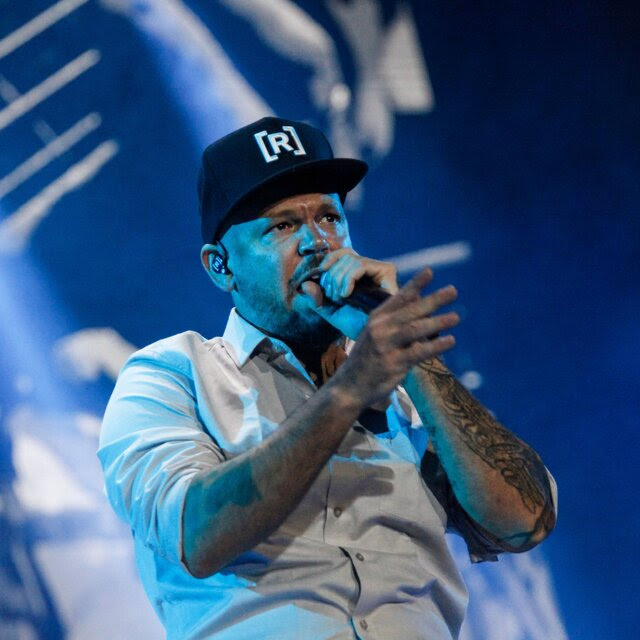 A man in a black ball cap with the letter R on it raps into a microphone onstage.