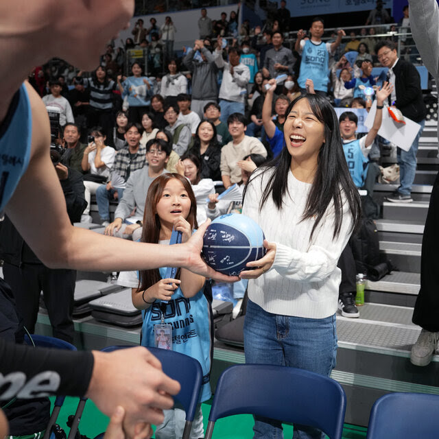 A male player in a sports uniform hands a blue basketball to a woman. Other fans who surround her are cheering.