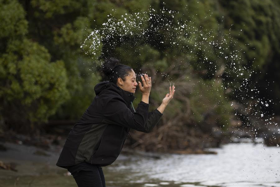 A woman stands in a river and uses her hands to throw a splash of water in the air.