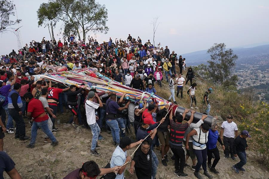 A crowd of men work together to carry a huge cross decorated with colorful ribbons. They are bringing the cross downhill. There is a crowd of people following the cross.