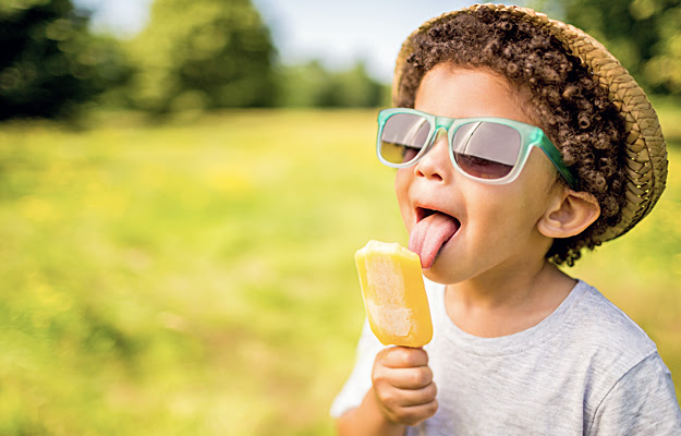 A young boy outside wearing sunglasses and a hat while eating a popsicle.