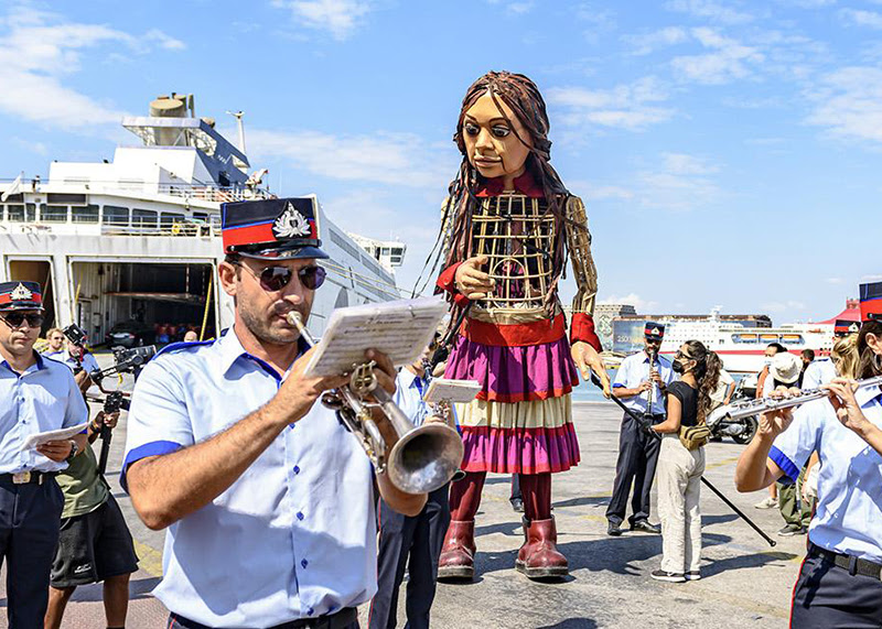 A photo of Little Amal, a 3.5m tall puppet. Amal is surrounded by a wind band playing their instruments as she arrives off a boat. The sky behind her is blue and the day is bright. 