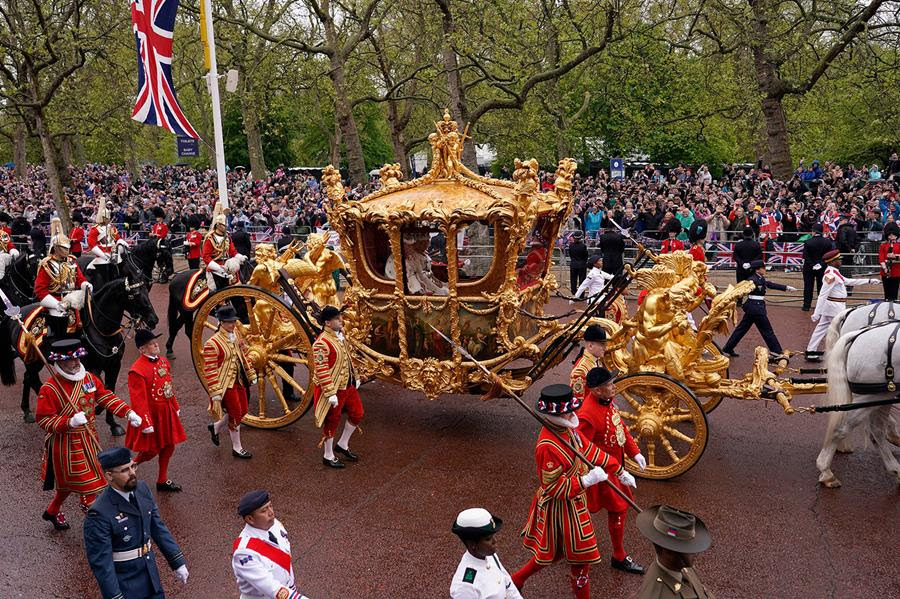 King Charles III and Queen Camila travel in the Gold State Coach following the coronation ceremony. The coach is being escorted by people in uniform and a crowd is gathered to watch.