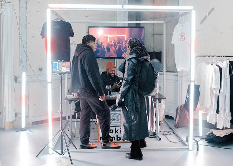 People interacting with a stall at the last Black Business Incubator Showcase. The stall is surrounded by bright white strip lights, with hand-designed t-shirts hanging from them.