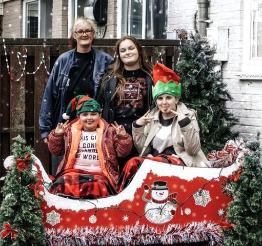 two adults and two children dressed in festive attire on a sleigh surrounded by christmas trees in a backyard