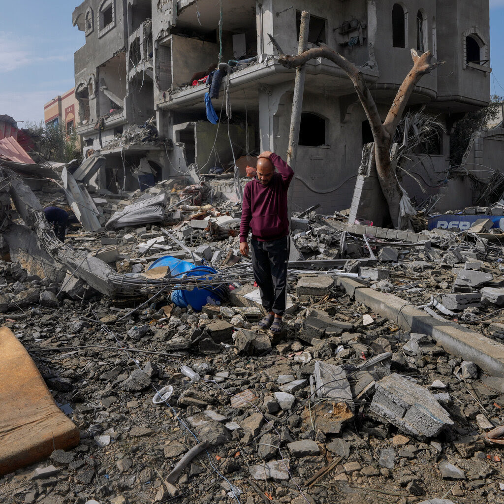 A man looks down at the ground, standing amid rubble in front of a badly damaged building.