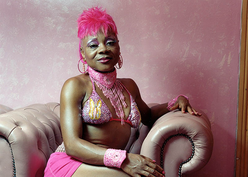 A woman with pink hair, pink bikini top, pink skirt and pink necklace and bracelets poses for the camera in a pink room, sat on a pink armchair.