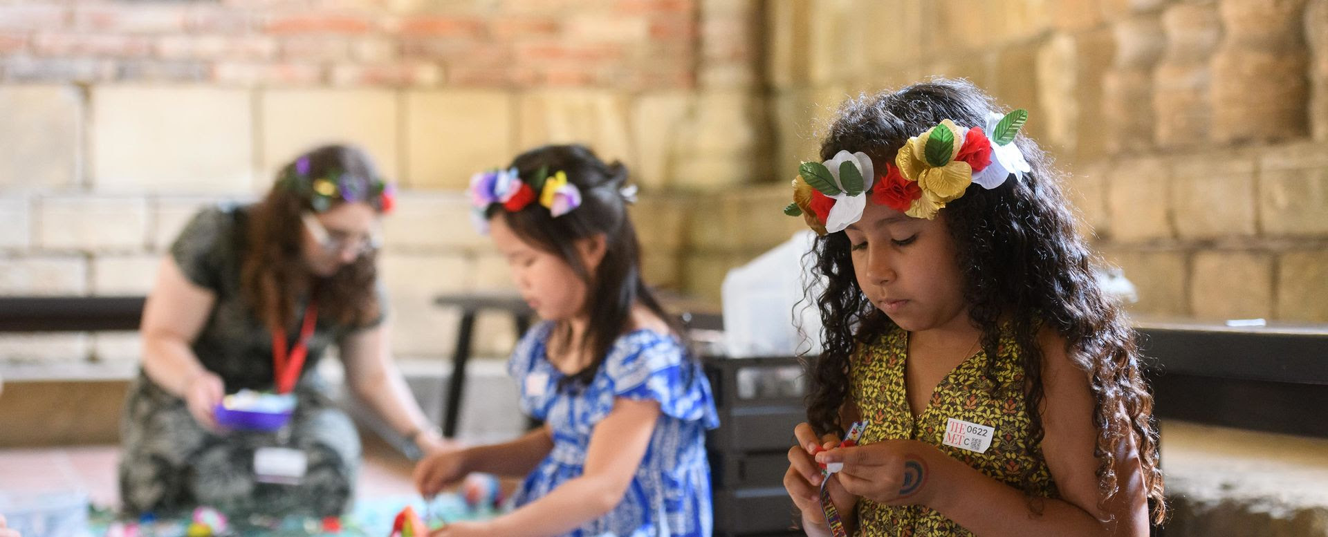 Duas meninas e uma mulher trabalham em artes e ofícios usando coroas de flores coloridas.