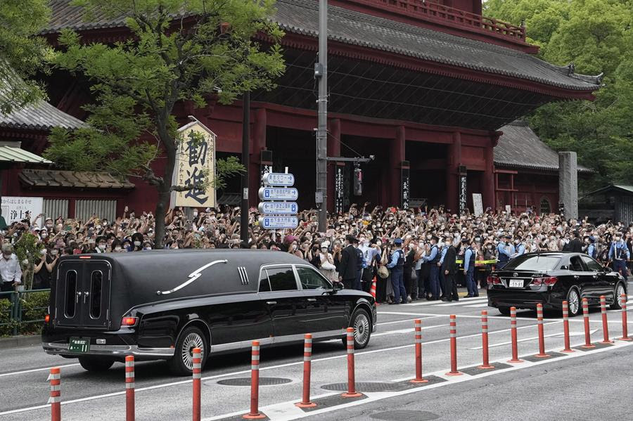 A black vehicle carrying the body of former Japanese Prime Minister Shinzo Abe leaves Zojiji temple. There are is a crowd of people at the side of the road watching the vehicle drive by.
