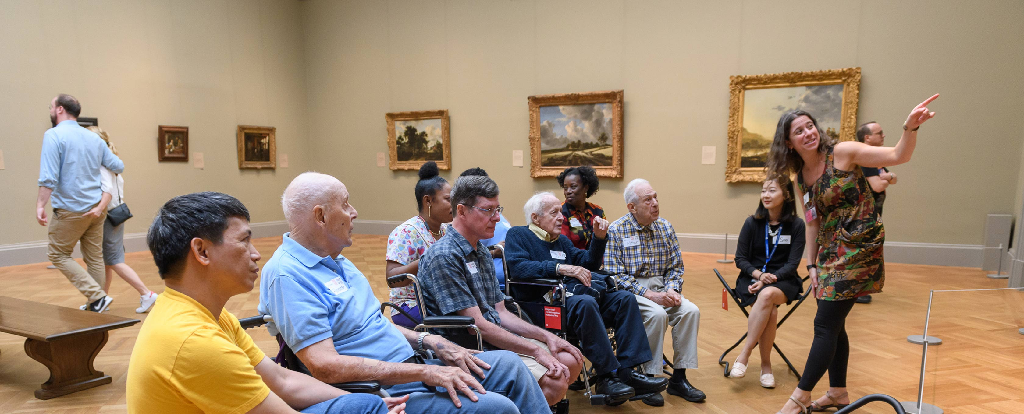 Um grupo está sentado em uma galeria ouvindo uma mulher falar sobre uma pintura.