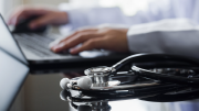 A medical professional uses a laptop. A stethoscope lies on a table in the foreground. 