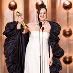 A smiling woman in a white sheath dress with a voluminous black coat holds a trophy aloft. 