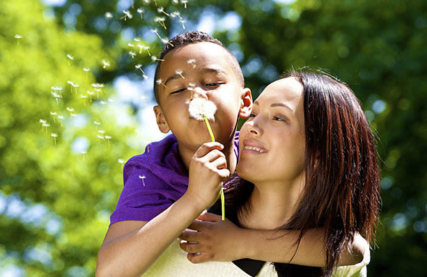 A little boy on his mother's back blowing seeds off of a dandelion.