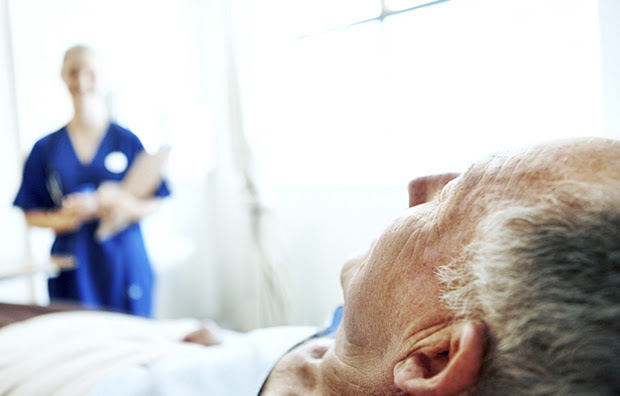 A senior man lying in a hospital bed with a nurse in the background.