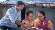 A father and mother read with their daughter in front of their home in Nepal. 