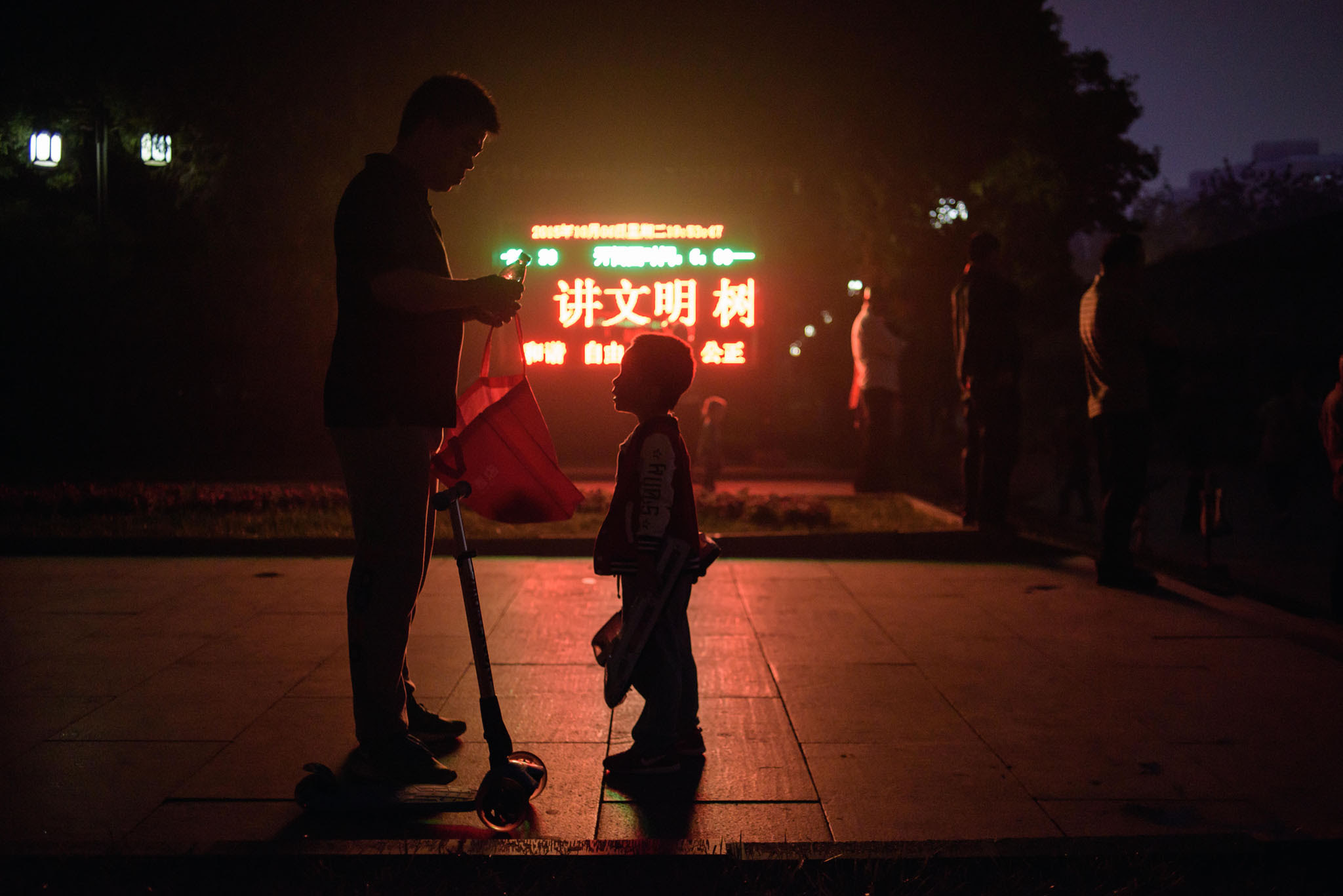 A man opens a drink to a child as they stand before a display board at the entrance to a park in Beijing