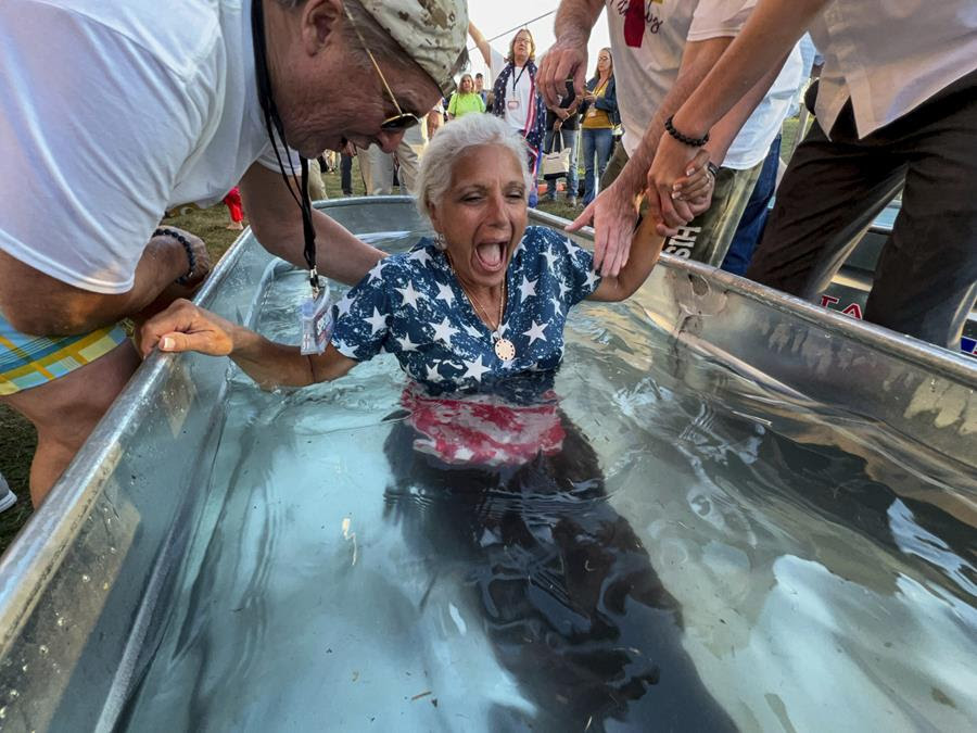 A woman is baptized in large metal tub. She is wearing a shirt patterened with the American flag and there are people gathered around her.