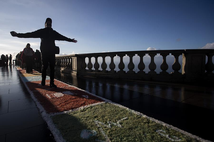 People walk on a rug made of colored salt. The person at the front of the procession has their arms outstretched and their head tipped towards the sky.
