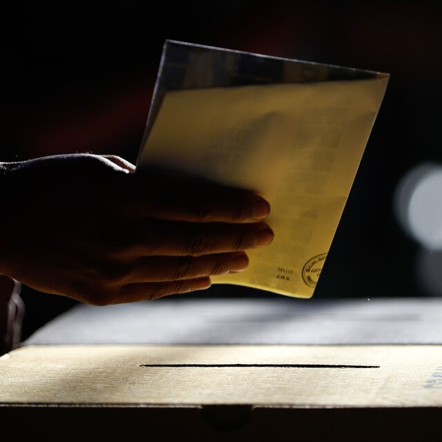 A photograph of a hand holding a folded ballot above a slot in a box.