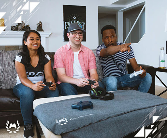 Three young adults sit on a couch at home playing Xbox One enthusiastically.
