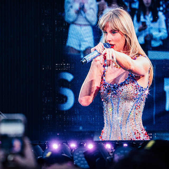 Taylor Swift onstage in concert wearing a glittery outfit and holding a microphone.