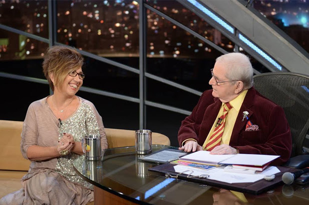 Descrição da imagem:Claudia Werneck durante entrevista com Jô Soares. Ela está sentada num sofá e olha sorridente para o apresentador. Ele está sentado numa cadeira, tateando o livro Sonhos do Dia, em braile, de Claudia Werneck, que está sobre a mesa, com mais três outros livros da autora e convidada.