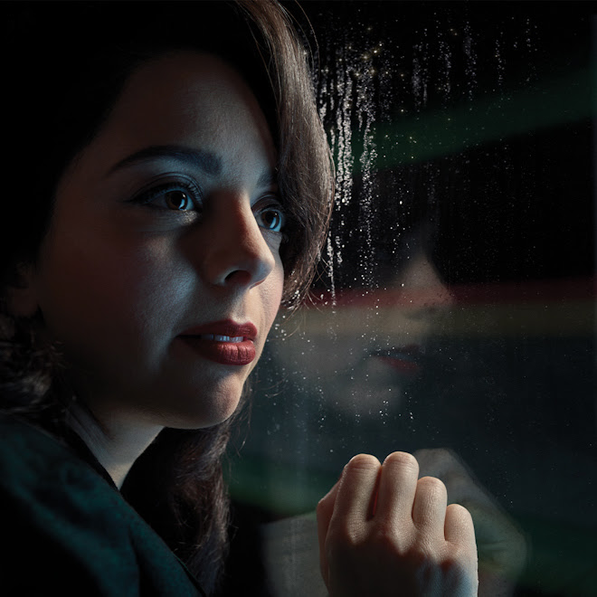 An image of a woman with brown hair, looking outside a window with a hand press against the glass.