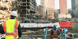 A construction worker in safety gear sits on a bucket, surveying the wreckage of the Twin Towers at Ground Zero in New York in September 2001. Behind him, an OSHA inspector in safety gear and a yellow OSHA vest looks at the scene.