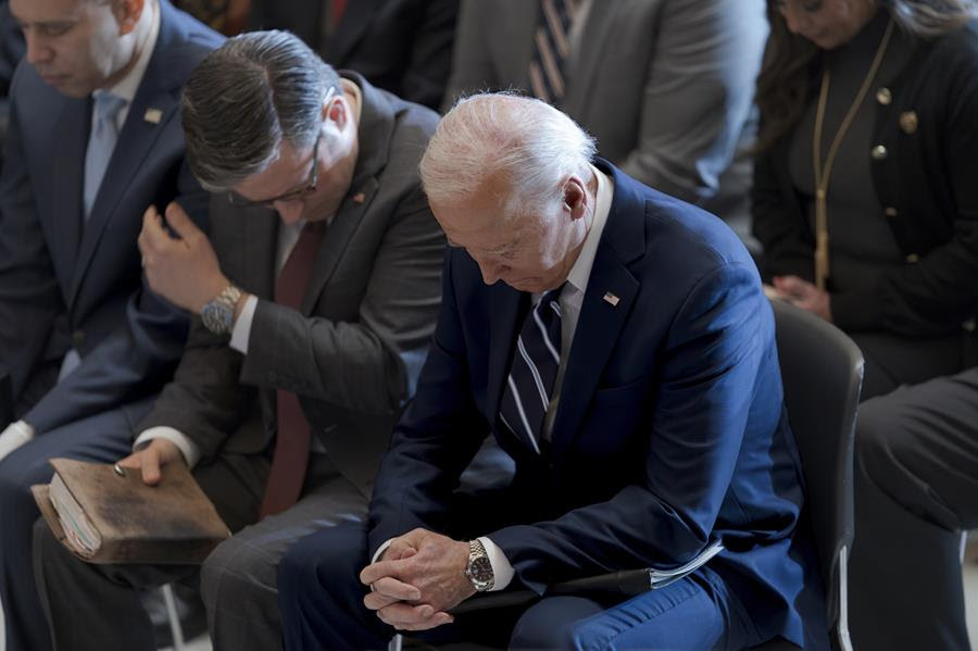 Joe Biden, sits beside Mike Johnson and Hakeem Jeffries during the National Prayer Breakfast. All three men have their heads bowed.