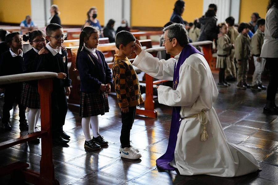 A Catholic priest kneels to mark a cross of ashes on a student's forehead. There are other students in line behind that student.