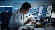 A woman in a lab coat leans over a desk, looking at something under a magnifying glass. 