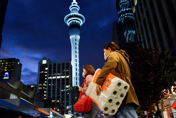 Two women wearing face masks and carrying a large pack of toilet paper walk past the illuminated Sky Tower in Auckland.