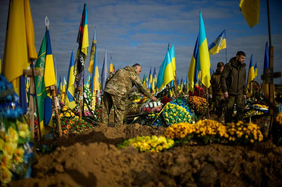 A Ukrainian serviceman places flowers on the grave of a recently killed fellow soldier in a cemetery. There are other graves surrounding him that also have flowers placed on them.