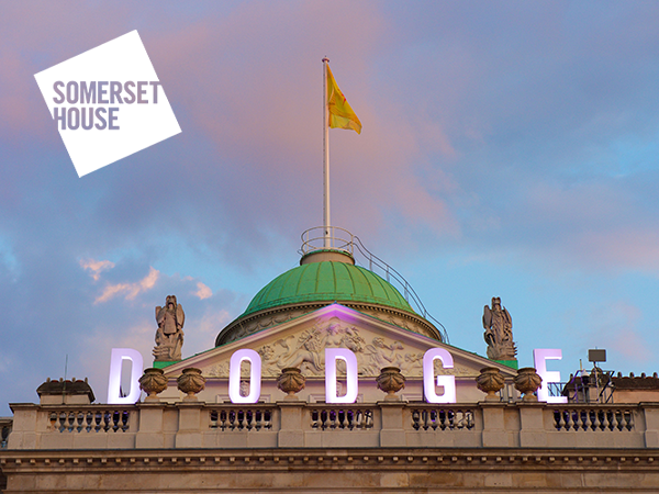 A photo of the South Wing of Somerset House. Pictured is the roof of the building with the letters DODGE positioned atop. The background is a blue sky, with light, pink clouds. The Future Producers yellow flag flies on top of the building.