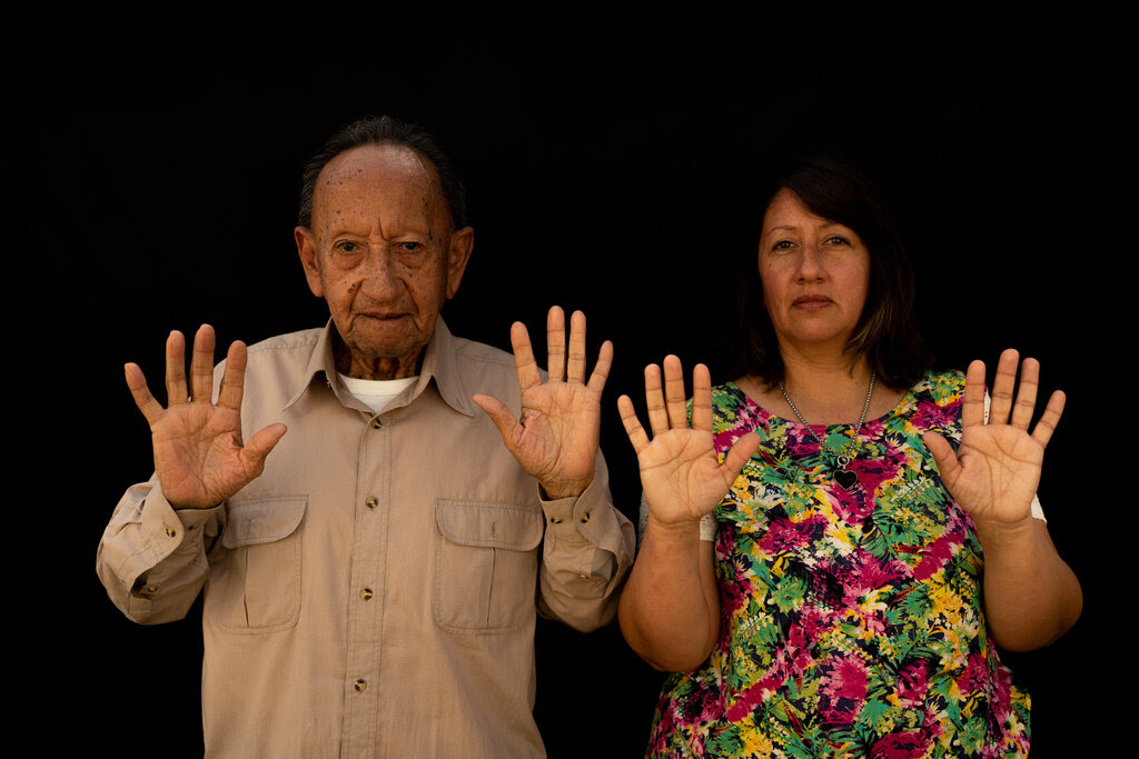 A man, left, wearing a tan shirt, and a woman in a colorful shirt hold their hands up in an Indigenous gesture.