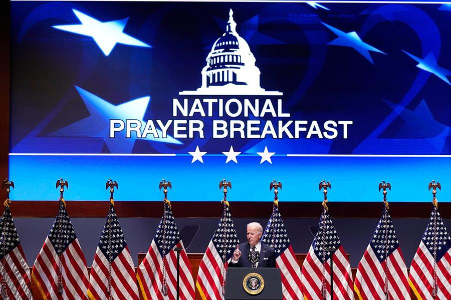 President Joe Biden speaks at the National Prayer Breakfast. There is a row of American flags behind him. The background is blue with the words "National Prayer Breakfast" in the center.