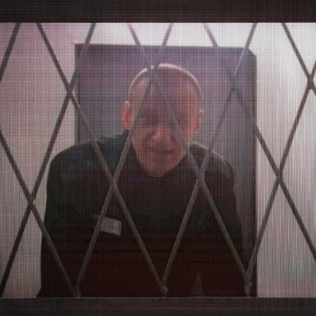 A still of Aleksei Navalny in black prison garb, seen through a metal grille.