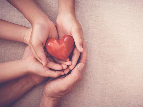 Photo looking down onto three pairs of hands holding a red love heart carefully.