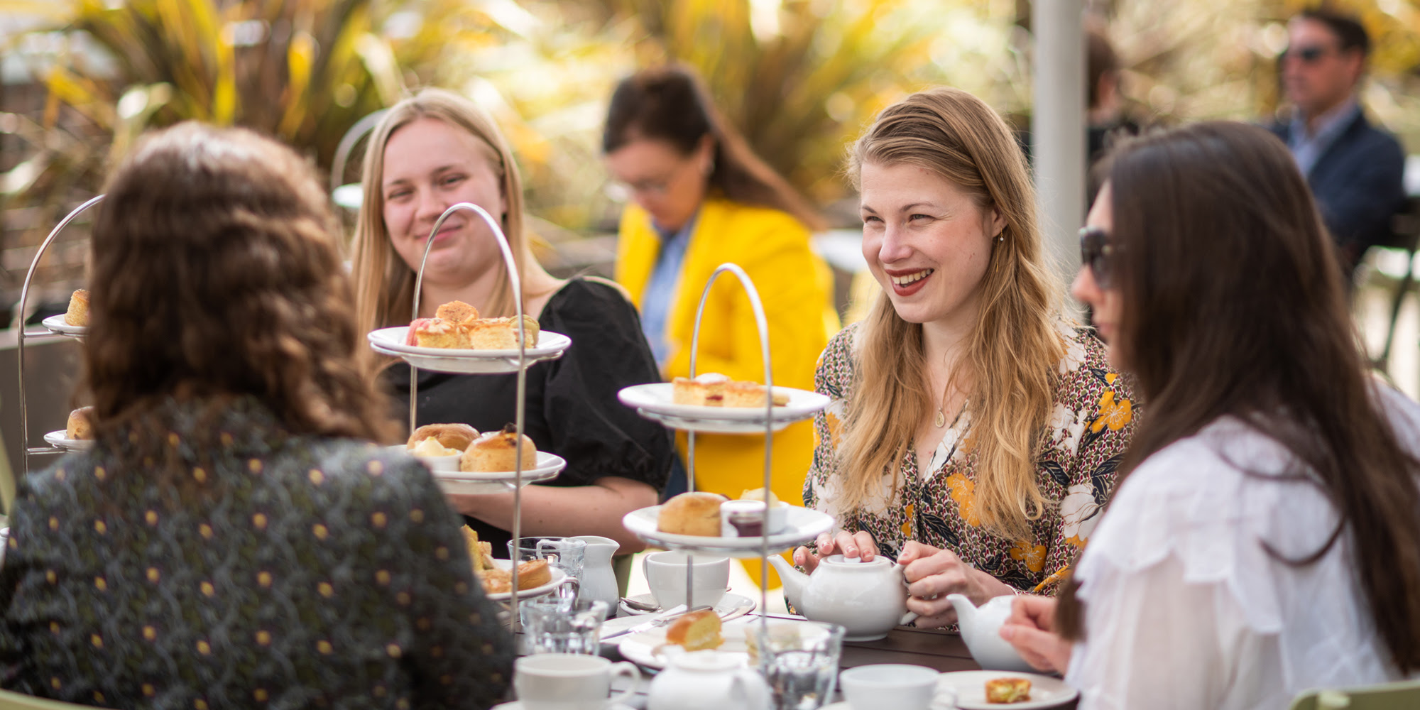 A group of women smile and enjoy traditional afternoon tea, with towers of cakes and scones in front of them