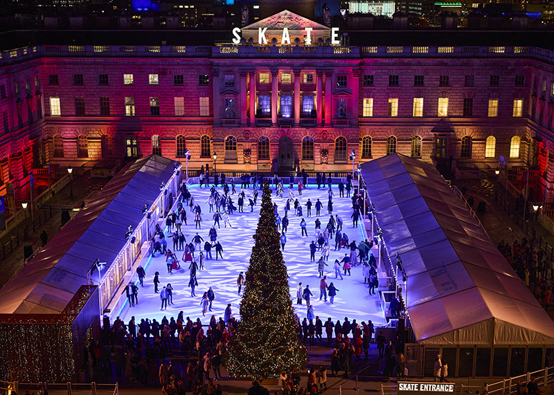 A photo of the ice rink at Somerset House. Ice skaters whizz across the ice, and light projections make pretty patterns on the ice. 