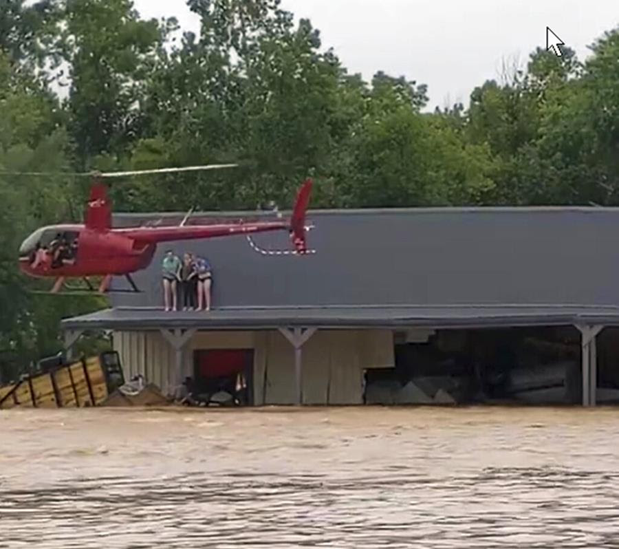 A red helicopter flies next to the roof of a building. There are people sheltering from muddy flood water on the roof.