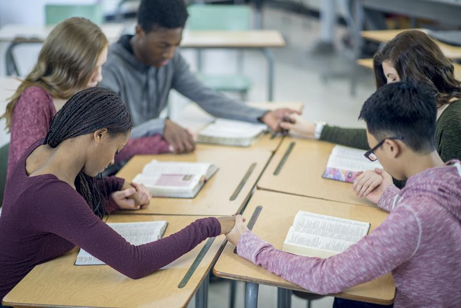 A diverse group of five teenagers hold hands, with heads bowed, in a classroom.