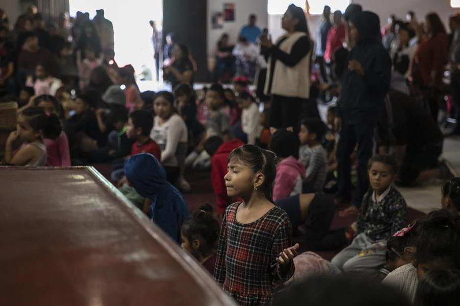 A young girl prays during a Christian service. There are many people in the background sitting on the floor or standing.