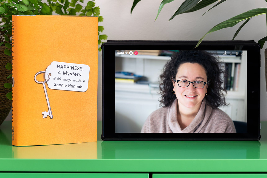 Photograph of a tablet standing on a green cabinet next to two houseplants and a book. The book has a bright orange cover with an illustration of a key and white label. The book titles appear on the key's label in black text  "Happiness, A Mystery & 66 attempts to solve it" by "Sophie Hannah". A photographic portrait of Sophie Hannah is on the tablet screen with video call icons "People", "Chat" and a red telephone icon.