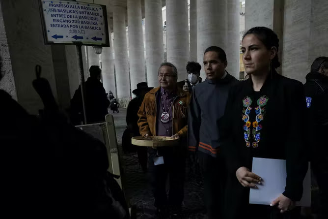 A woman in a black shirt holding a document stands in front of two men in St. Peter's Square at the Vatican.