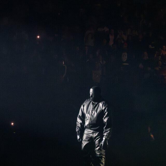 A man dressed all in black leather, wearing a full mask, stands with his arms at his sides, surrounded by darkness.